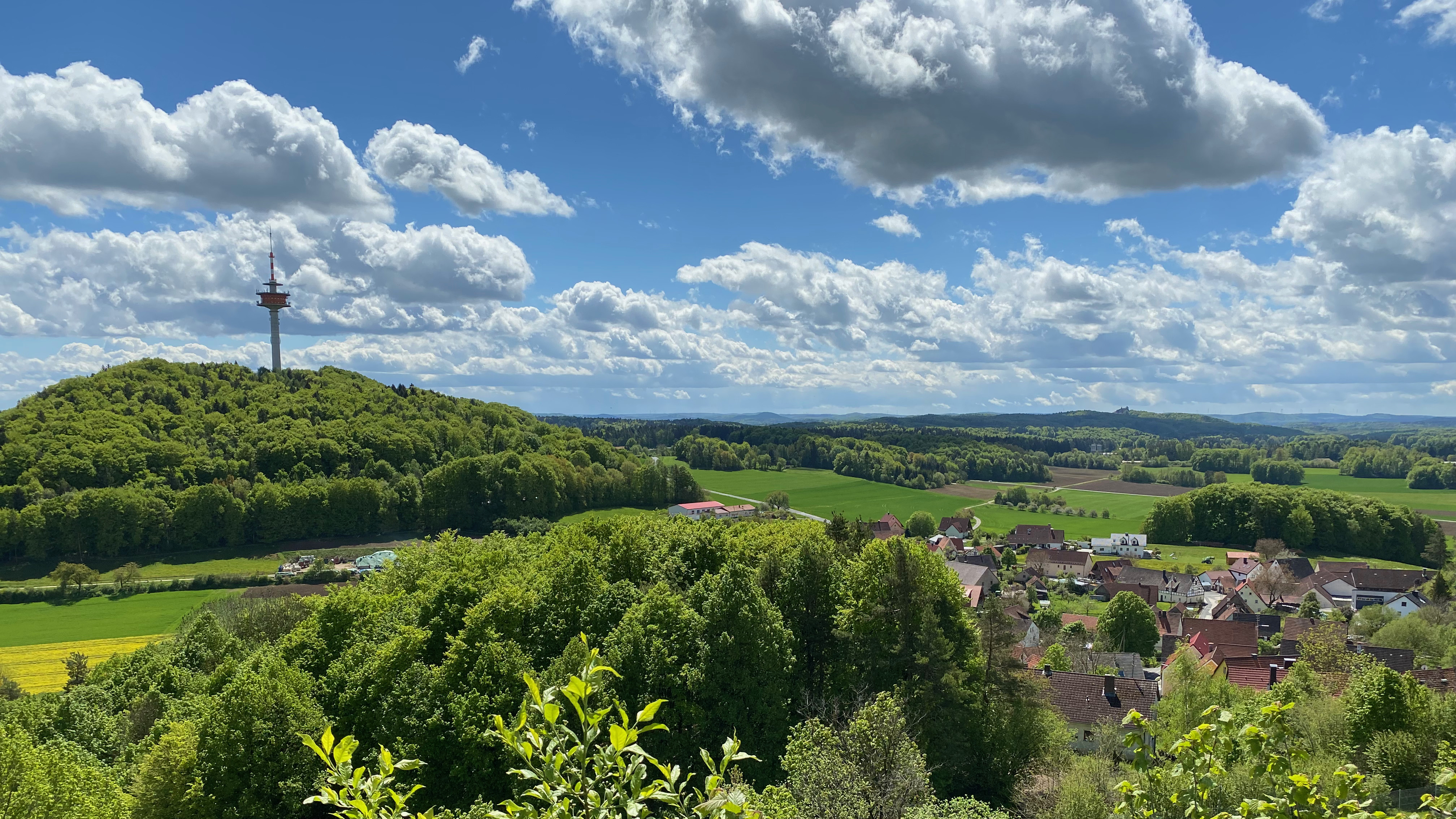 Landschaft mit Hügeln, Fernsehturm und Dorf unter blauem Himmel mit Wolken.